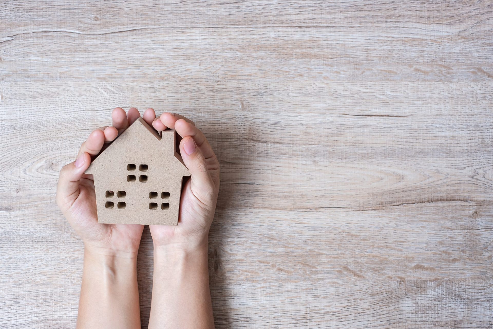 Two hands hold a small cardboard cutout of a house with window shapes, placed on a light wooden surface—symbolizing the comfort and security of a lifetime promise.