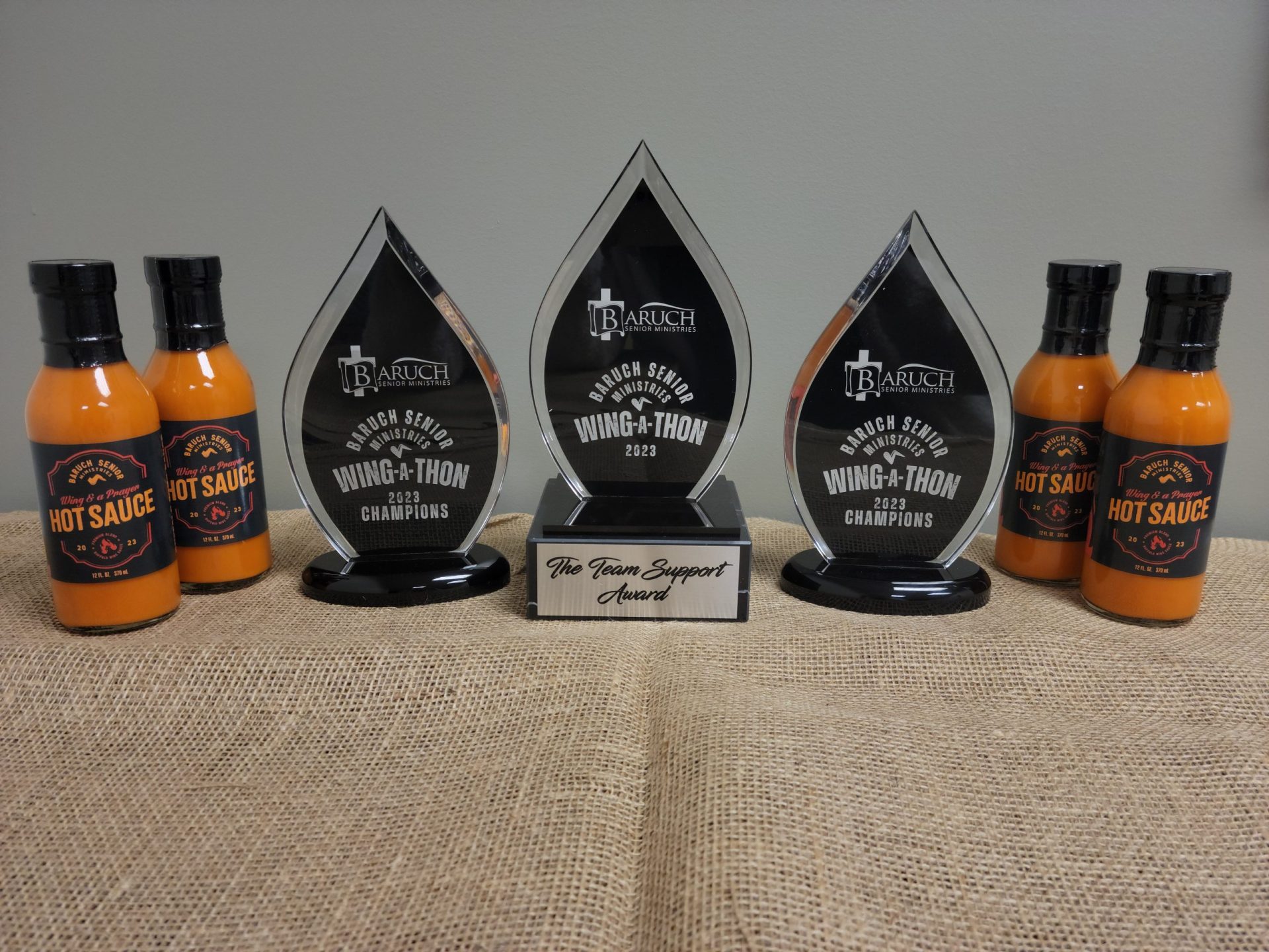 Three crystal trophies labeled "Wing-a-Thon 2023 Champions" and "The Team Support Award" sit on burlap, flanked by six bottles of hot sauce—prizes from Baruch Senior Ministries’ virtual wing-eating fundraiser.