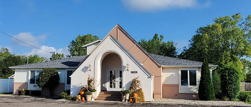 Single-story building with a central arched entrance, white and peach exterior, small front steps, and fall decorations including pumpkins and cornstalks by the doorway. Single-story building with a central arched entrance, white and peach exterior, small front steps, and fall decorations including pumpkins and cornstalks by the doorway.