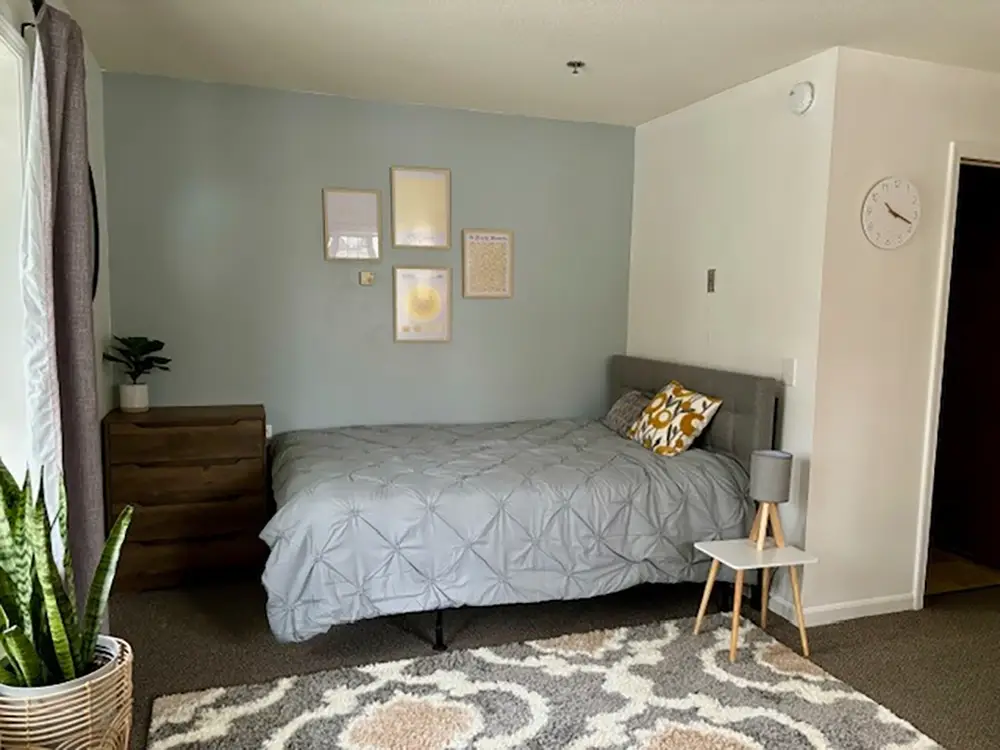 A neatly made bed with gray bedding sits against a light blue accent wall decorated with framed pictures; a dresser, small side table, and clock are also visible in the room. A neatly made bed with gray bedding sits against a light blue accent wall decorated with framed pictures; a dresser, small side table, and clock are also visible in the room.