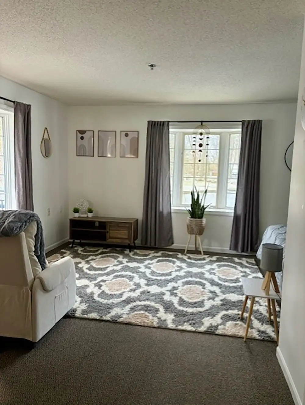 A living room with a patterned rug, two windows with gray curtains, a white armchair, a TV stand, a plant, and minimalist wall art. A living room with a patterned rug, two windows with gray curtains, a white armchair, a TV stand, a plant, and minimalist wall art.