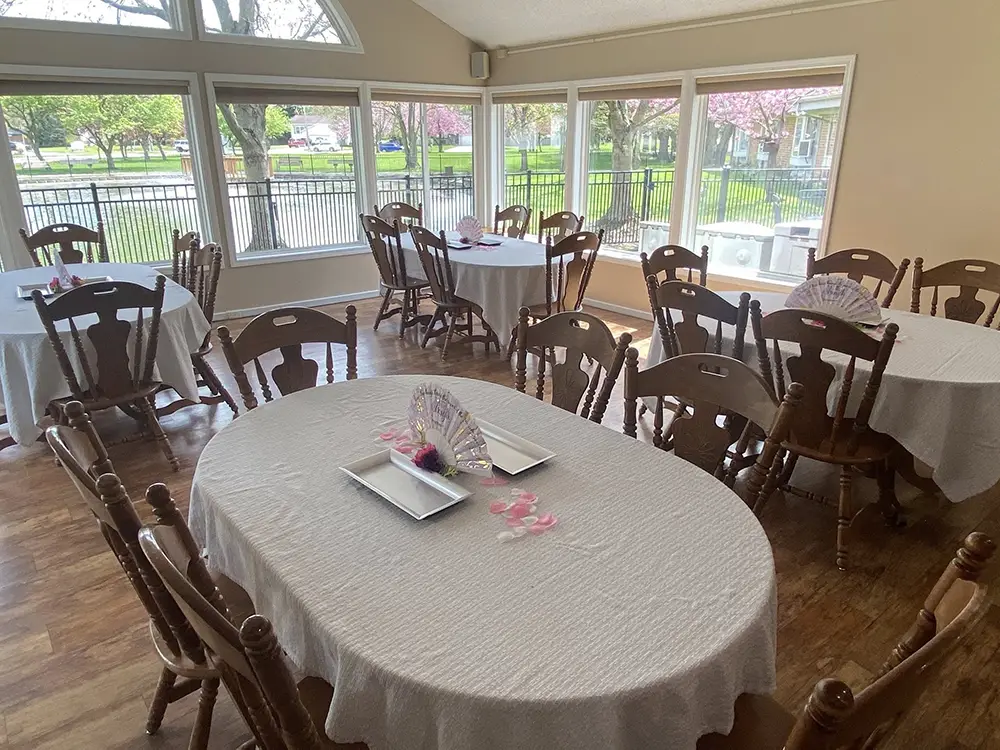 Room with large windows, several round tables covered in white cloths, wooden chairs, and pink and white decorative centerpieces on the tables. Room with large windows, several round tables covered in white cloths, wooden chairs, and pink and white decorative centerpieces on the tables.