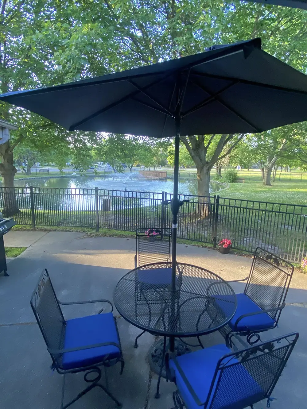 A round metal patio table with four blue-cushioned chairs and a large umbrella overlooks a fenced yard with trees and a pond in the background. A round metal patio table with four blue-cushioned chairs and a large umbrella overlooks a fenced yard with trees and a pond in the background.