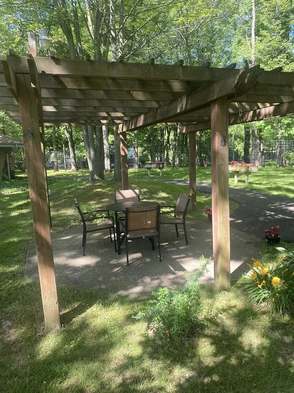 Wooden pergola with table and four chairs on a concrete patio in a grassy yard, surrounded by trees, flowers, and sunlight. Wooden pergola with table and four chairs on a concrete patio in a grassy yard, surrounded by trees, flowers, and sunlight.