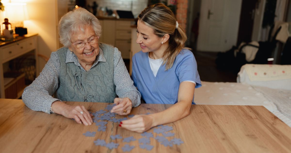 An elderly woman and a caregiver in scrubs sit at a table, smiling and working together on assembling a jigsaw puzzle—one of the enriching dementia care activities often featured during a memory care community tour.