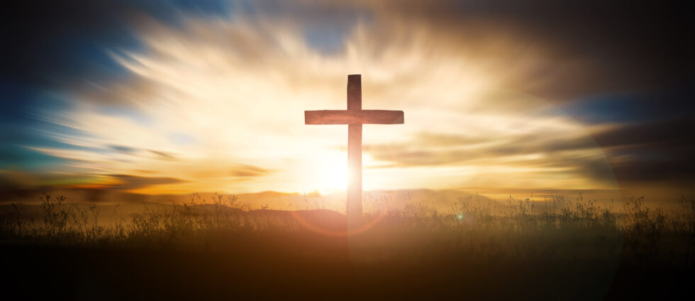 A wooden cross stands on a hill with the sun setting or rising behind it, casting light through a cloudy sky—a powerful scene for Holy Week reflection.