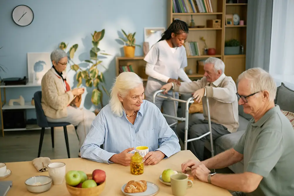 Five seniors are in a common room; two sit at a table with drinks and fruit, one knits, one reads, and a caregiver assists a man with a walker in the background.