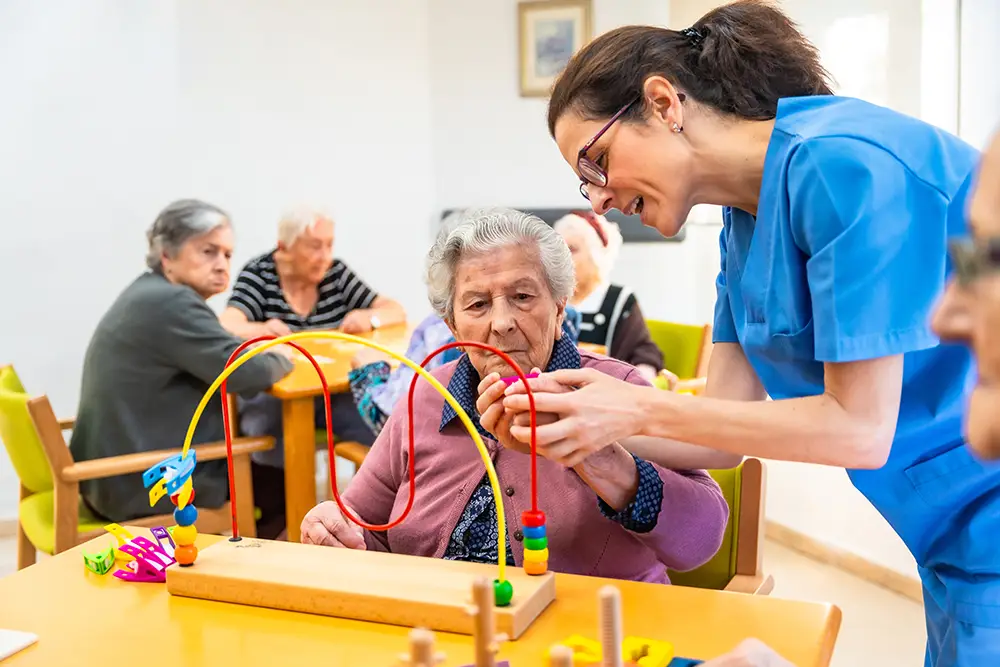 A caregiver assists an elderly woman with a bead maze activity at a care facility, while other seniors sit at a nearby table.