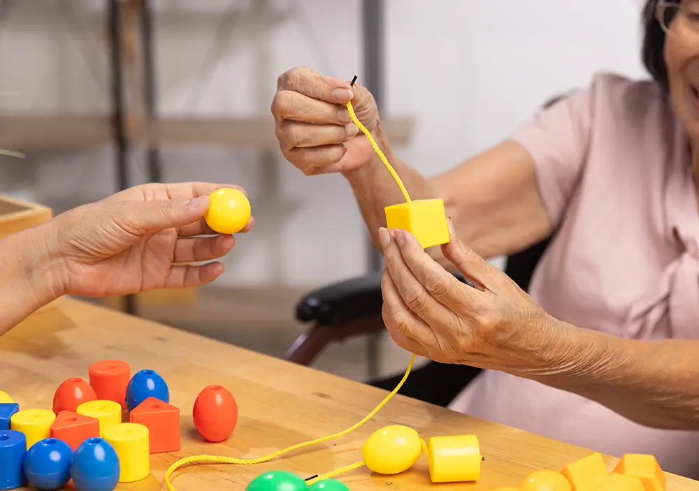 Two people use their hands to thread yellow plastic beads and shapes onto a string at a wooden table with colorful bead pieces scattered around.