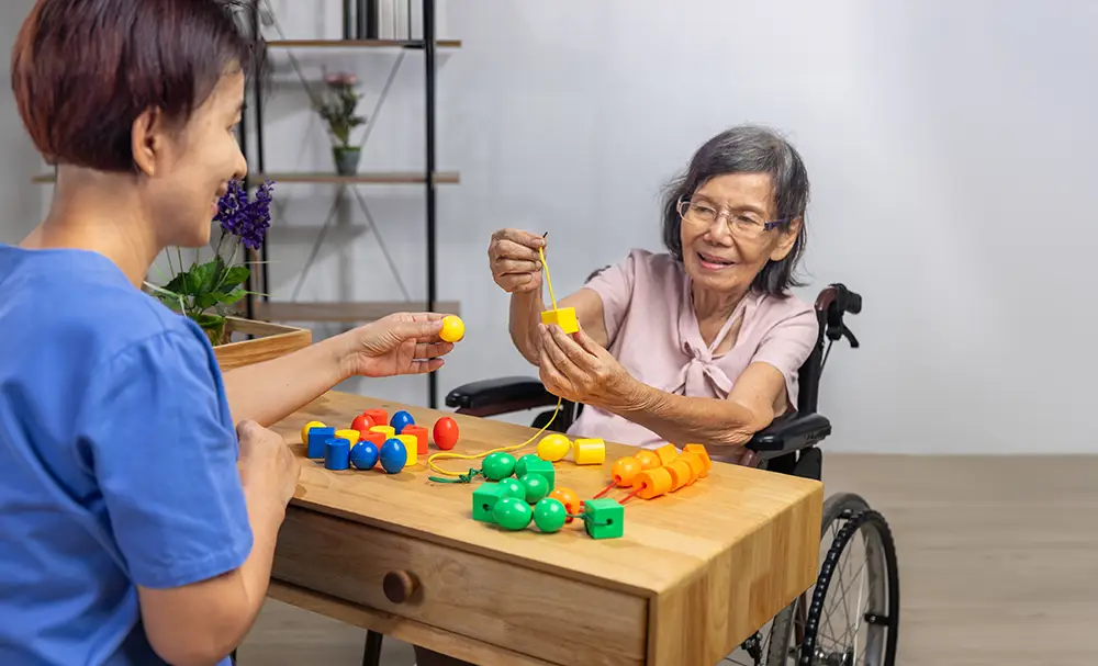 An elderly woman in a wheelchair strings colorful beads with the help of a caregiver at a wooden table during a therapy session.