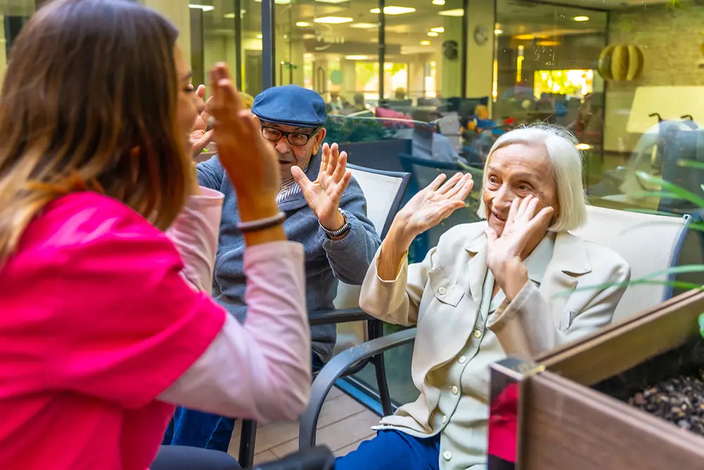 A caregiver and two elderly people sit outdoors, smiling and raising their hands in a playful gesture at a senior living facility.