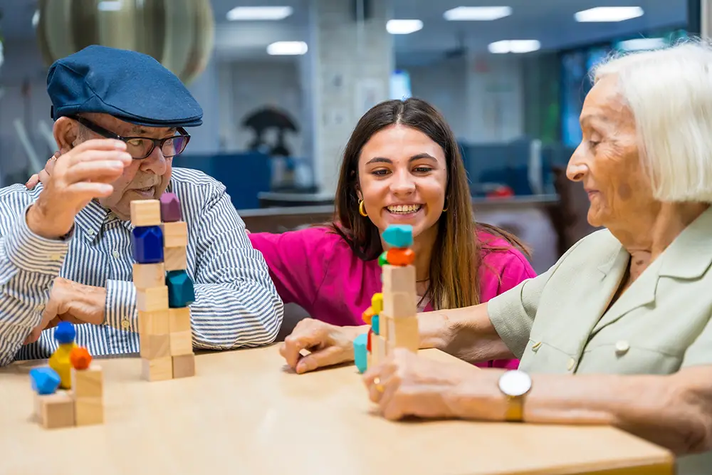 Two elderly people and a caregiver sit at a table, smiling and stacking colorful blocks in a well-lit communal space.
