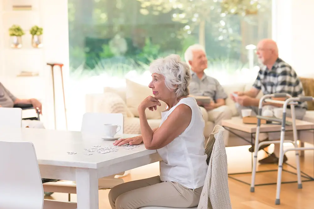 An older woman sits at a table working on a puzzle, while three other elderly people talk and relax in a bright, sunlit common room.