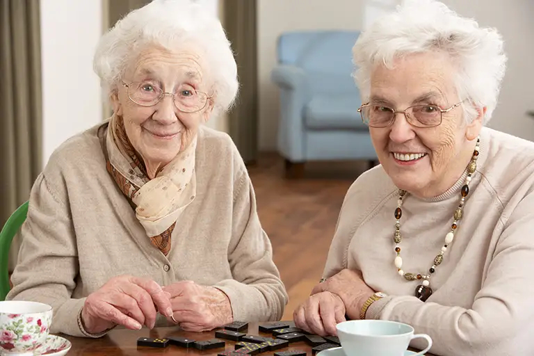 Two elderly women sit at a table, smiling, while playing dominoes with tea cups nearby in a cozy living room setting.