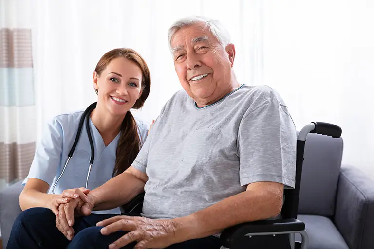 A nurse sits beside an elderly man in a wheelchair, smiling and holding his hand in a bright, indoor setting.