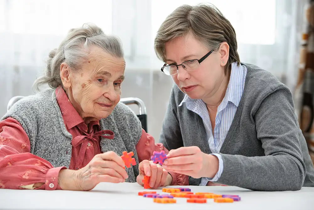 An elderly woman and a younger woman sit at a table working together to assemble a colorful jigsaw puzzle.