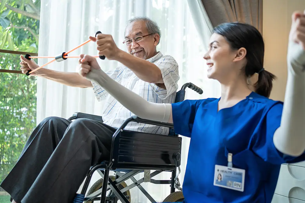 Elderly man in a wheelchair using a resistance band for exercise with assistance from a female healthcare professional in blue scrubs. Elderly man in a wheelchair using a resistance band for exercise with assistance from a female healthcare professional in blue scrubs.