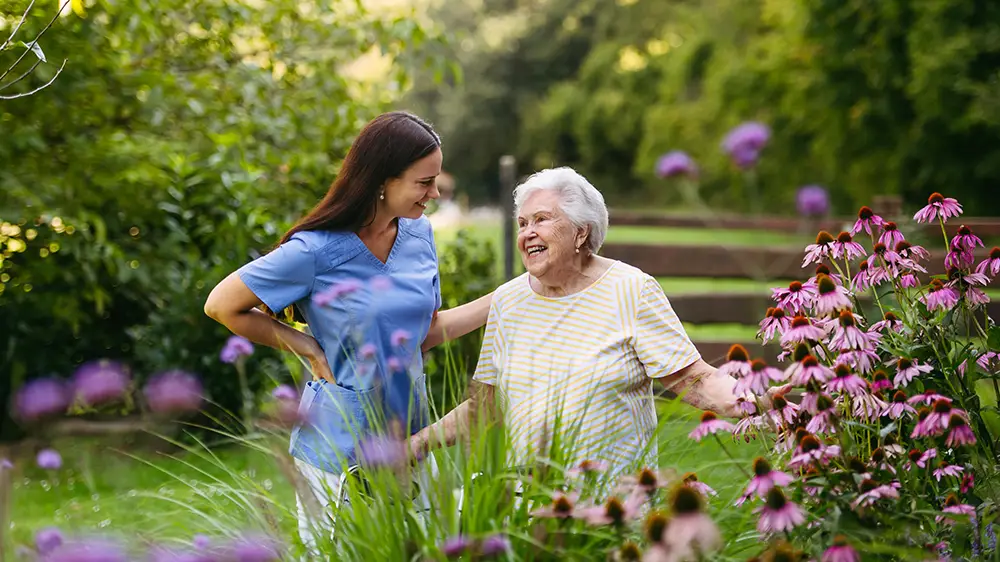 A young woman and an elderly woman stand together in a garden, smiling and surrounded by pink flowers and greenery on a sunny day.