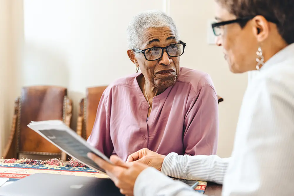 Two women sit at a table having a conversation; one holds documents while the other listens attentively, both appearing engaged in discussion.