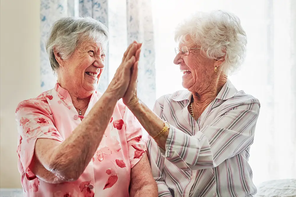 Two elderly women sit side by side, smiling and giving each other a high-five in a bright room with floral curtains.