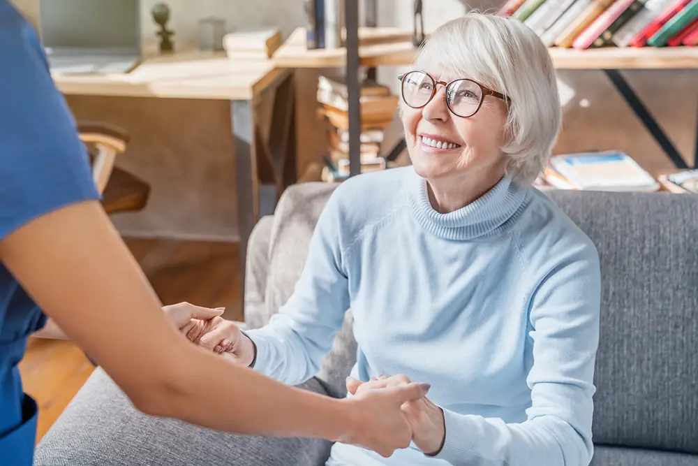 An older woman with short gray hair and glasses sits on a sofa, smiling and holding hands with a person in blue scrubs.