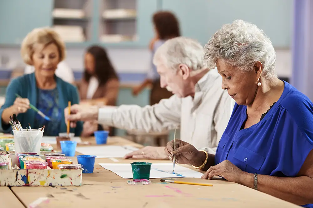 A group of older adults sit at a table painting with brushes and watercolors in an art classroom.