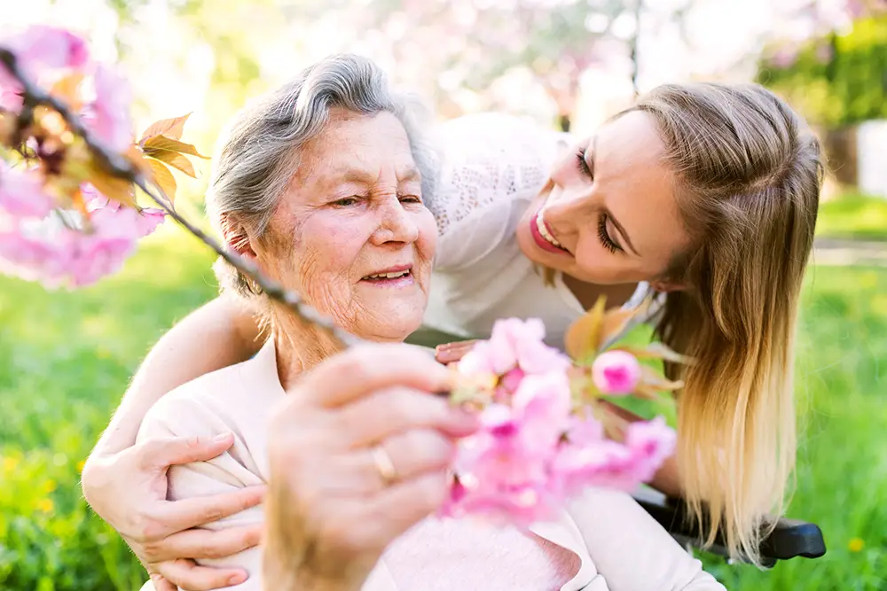 An older woman in a wheelchair holds pink flowers while a younger woman smiles and embraces her outdoors in a park.