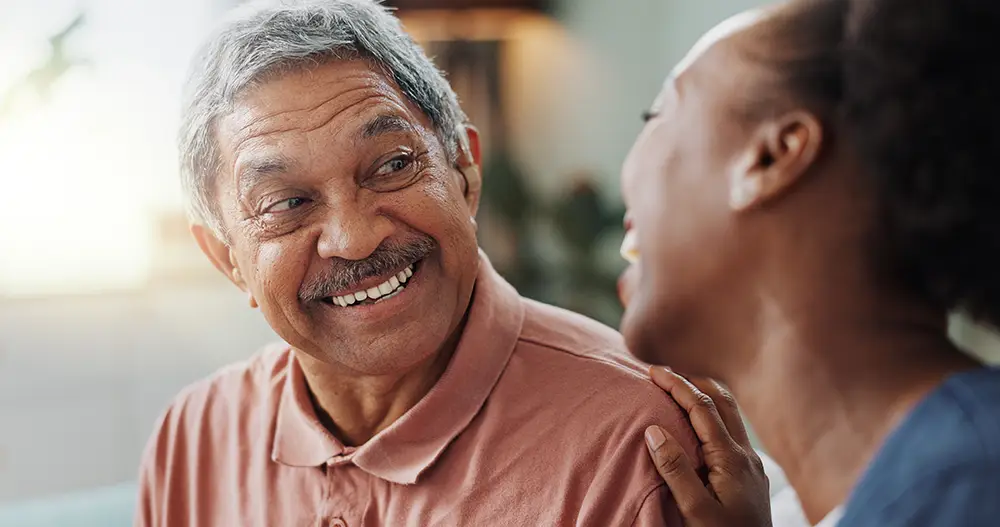 An older man in a polo shirt smiles while looking at a woman who is laughing, with her hand resting on his shoulder.