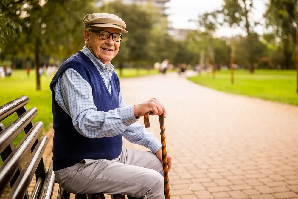 An elderly man wearing glasses, a cap, and a blue sweater vest sits on a park bench, holding a wooden cane, with a walkway and green trees in the background.