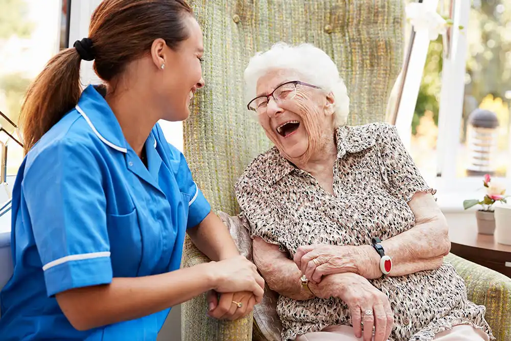 A nurse in blue scrubs smiles and holds hands with an elderly woman in a chair, who is laughing.