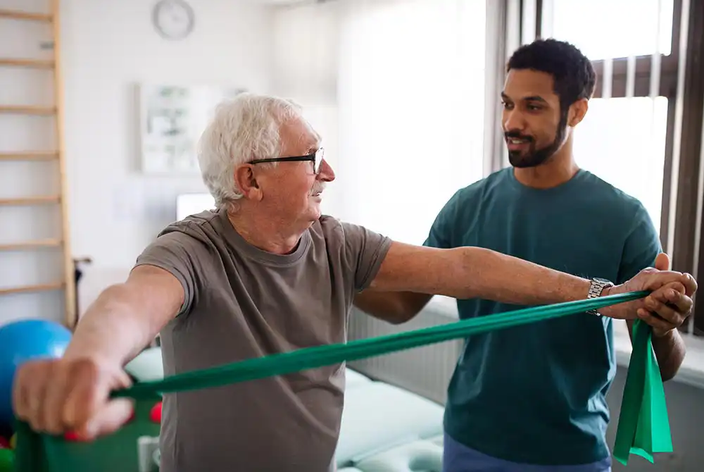 Older man doing arm exercises with a green resistance band while a younger man assists him in a brightly lit room.
