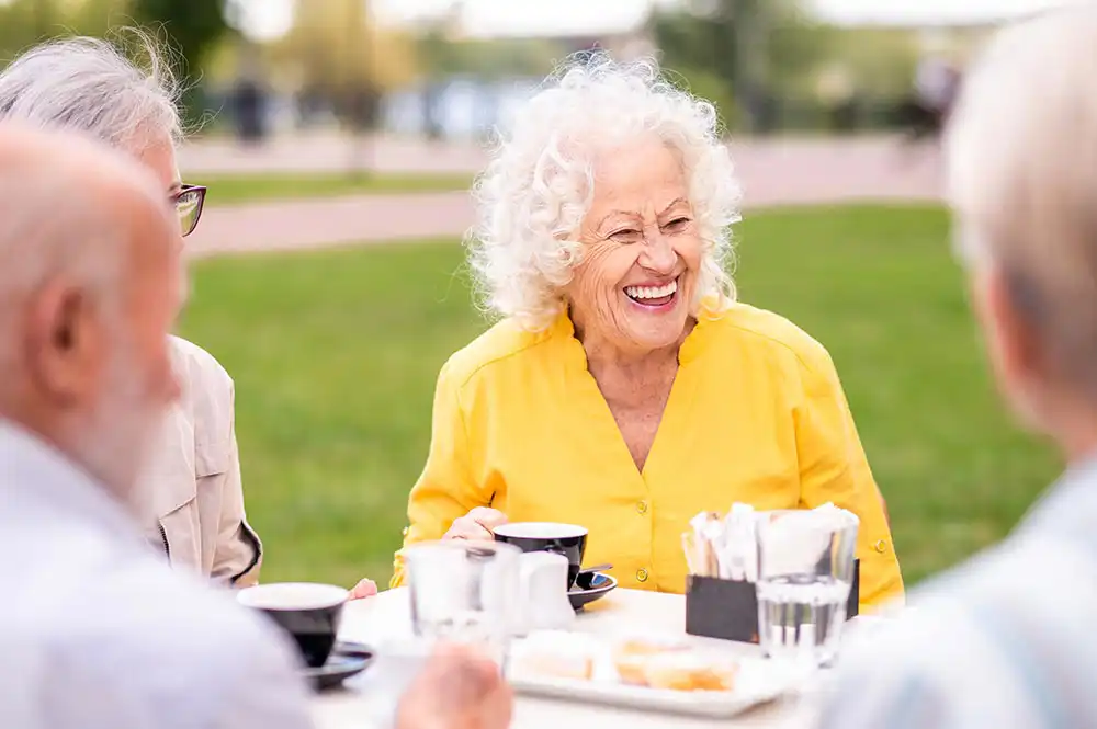 An older woman in a yellow shirt smiles while sitting outdoors at a table with three others, drinking coffee and chatting in a park setting.