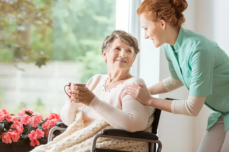 A caregiver in a green uniform smiles and talks to an elderly woman in a wheelchair, who is holding a mug and covered with a knit blanket near a window with pink flowers.