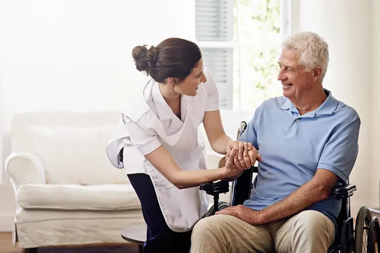 A caregiver assists an older man in a wheelchair, smiling at him while holding his hand in a bright living room.