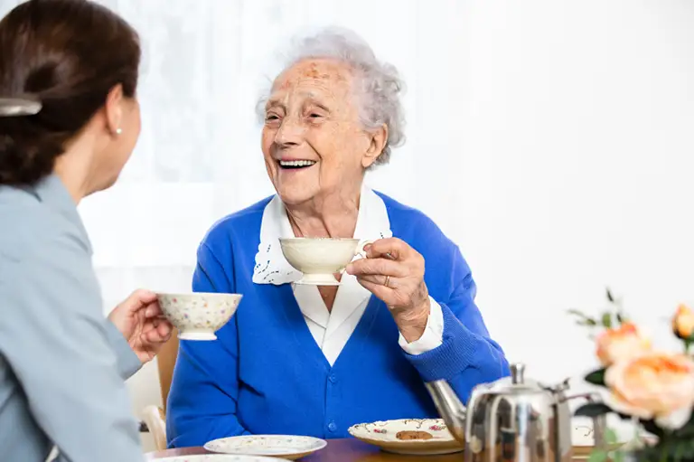 An elderly woman in a blue cardigan smiles and holds a teacup while sitting at a table and talking with another woman. A teapot and flowers are on the table.