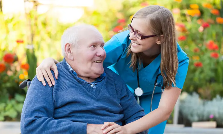 A nurse in blue scrubs smiles and embraces an elderly man in a blue sweater sitting in a wheelchair, outdoors with flowers in the background.