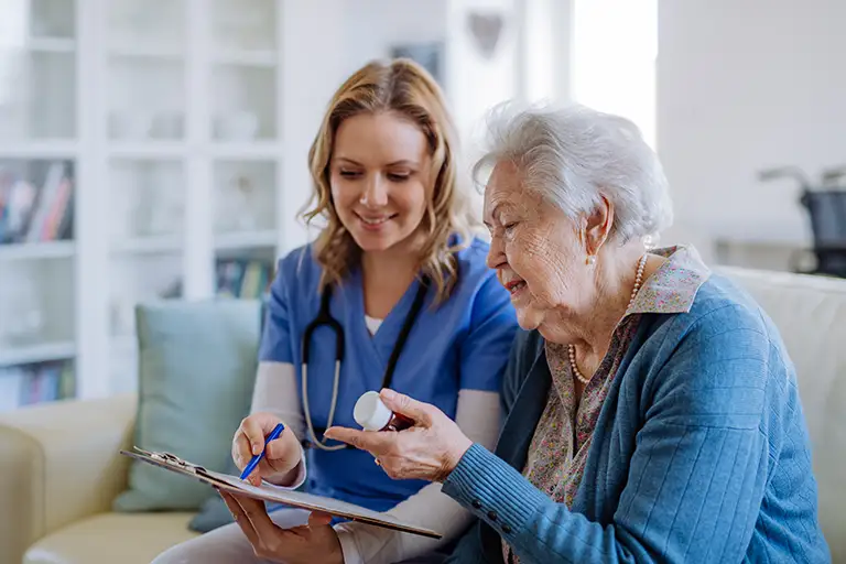 A nurse with a clipboard assists an elderly woman who is holding a pill bottle, both seated on a couch in a bright room.