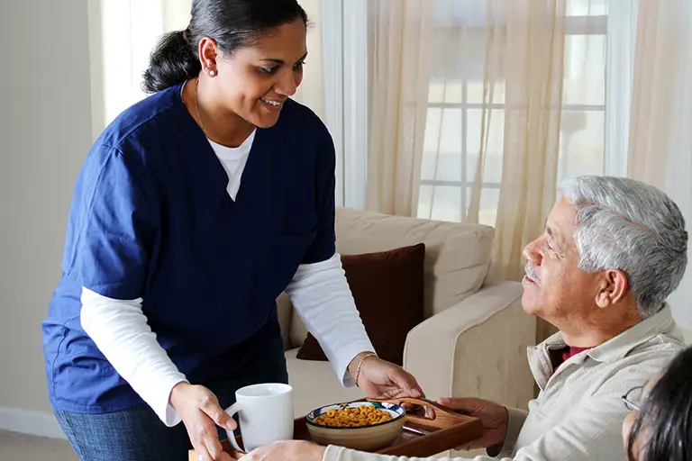 A caregiver in blue scrubs hands a meal tray with a bowl of food and a mug to an older man seated in a living room.