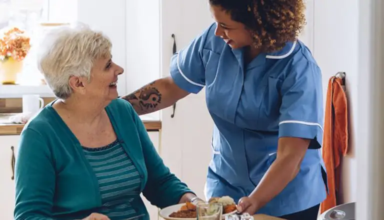 A caregiver in a blue uniform serves a meal to an older woman seated at a table in a kitchen. Both are smiling.