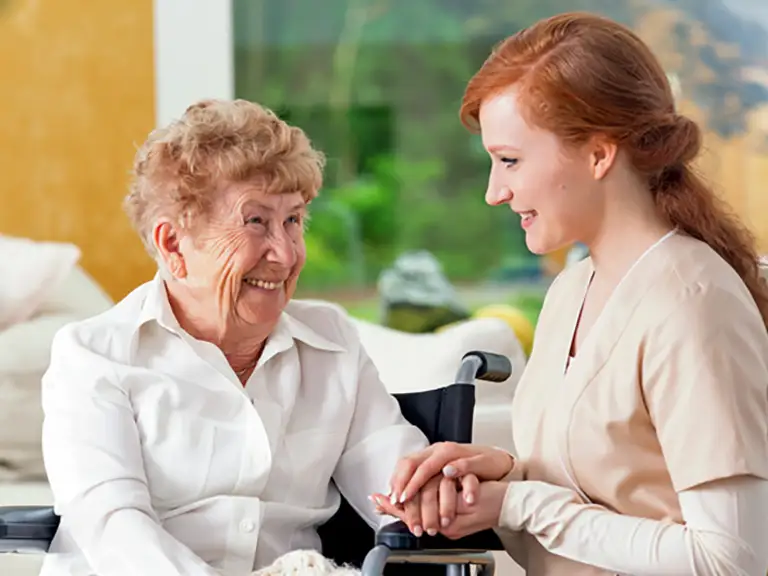 An elderly woman in a wheelchair smiles while holding hands with a young caregiver, who is also smiling, indoors.