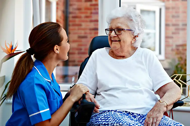 A nurse in blue scrubs sits beside an elderly woman in a wheelchair, holding her hand and smiling, in a bright indoor setting.
