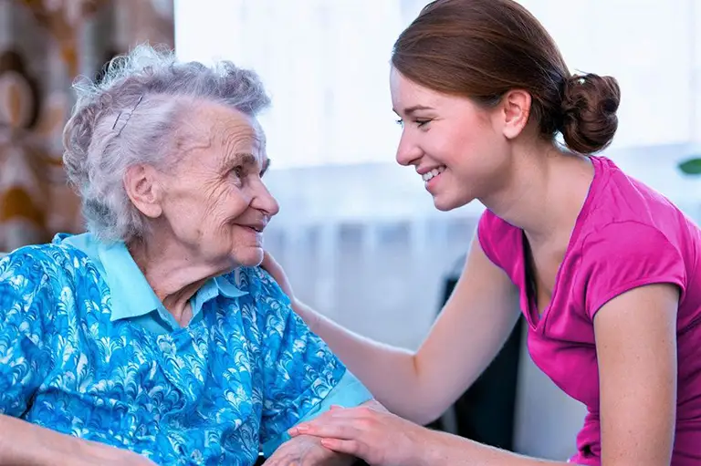 A young woman in a pink shirt smiles and holds hands with an elderly woman in a blue patterned blouse, sitting together indoors.