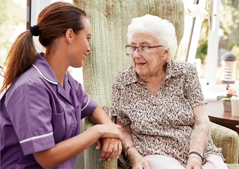 A caregiver in a purple uniform talks with an elderly woman sitting in a chair, both smiling in a well-lit room.