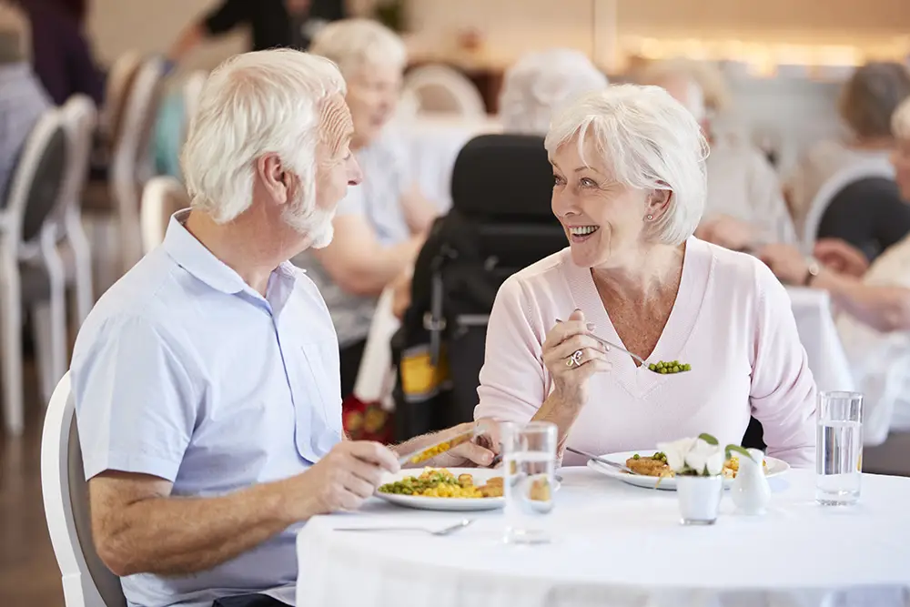 Two older adults sit at a table eating a meal and smiling at each other in a brightly lit communal dining area.