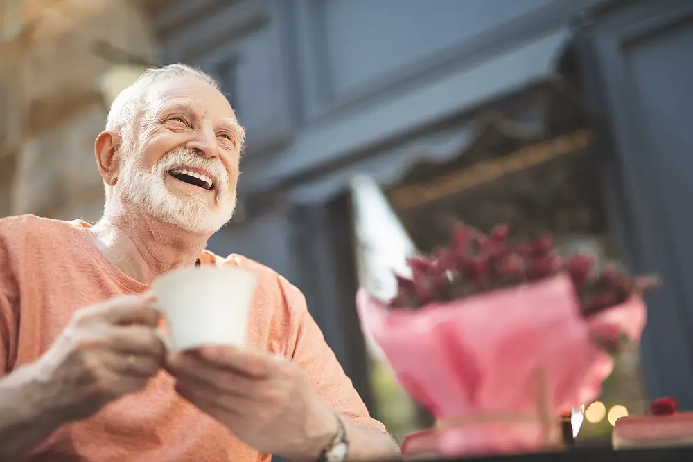 Older man with white hair and beard smiling and holding a cup of coffee, sitting at a table with a bouquet of flowers wrapped in pink paper.