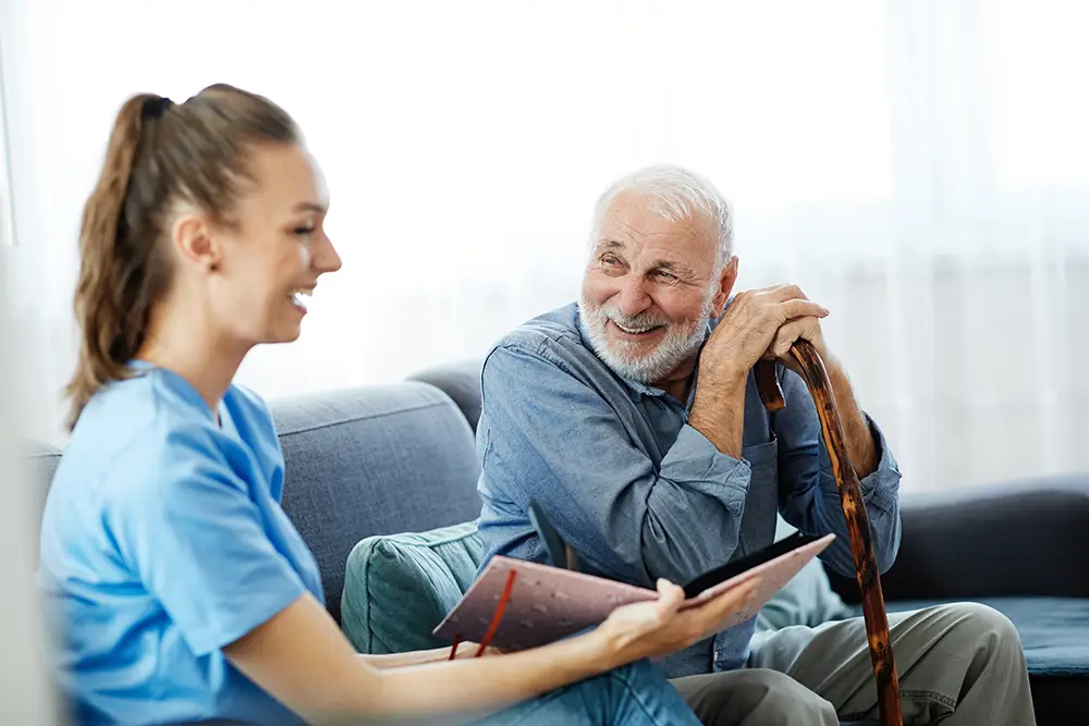 A young woman in scrubs shows a book to an older man with a cane, both smiling while sitting on a couch in a bright room.