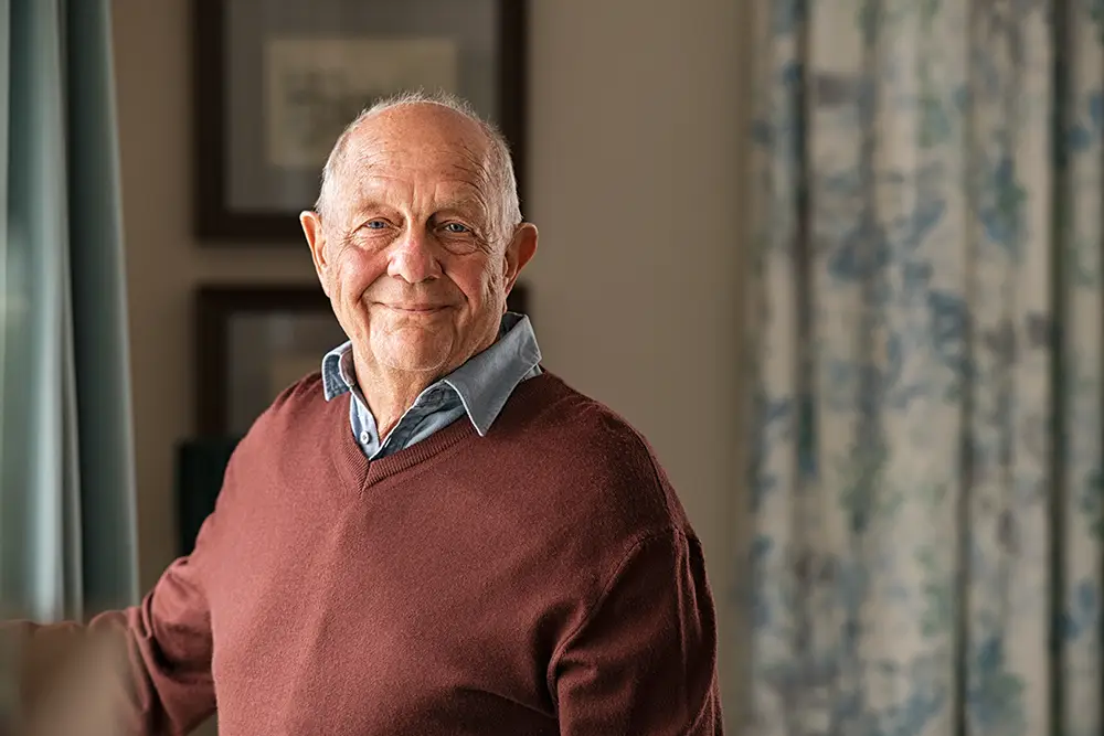 Older man with short gray hair wearing a maroon sweater over a collared shirt, standing indoors and smiling at the camera.