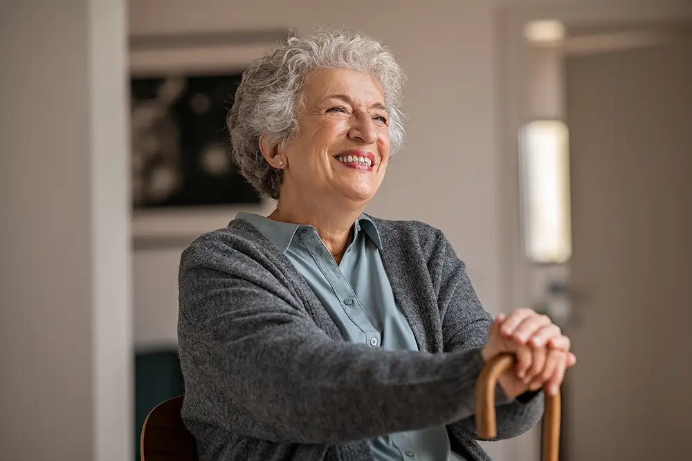 Smiling older woman with gray hair sits indoors, wearing a blue shirt and gray cardigan, resting her hands on a wooden cane.