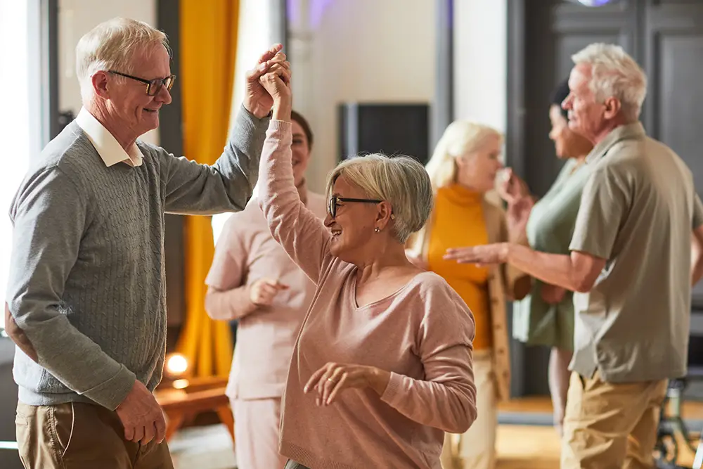 Older adults dance and socialize in a well-lit room, smiling and enjoying themselves in a casual, friendly atmosphere.