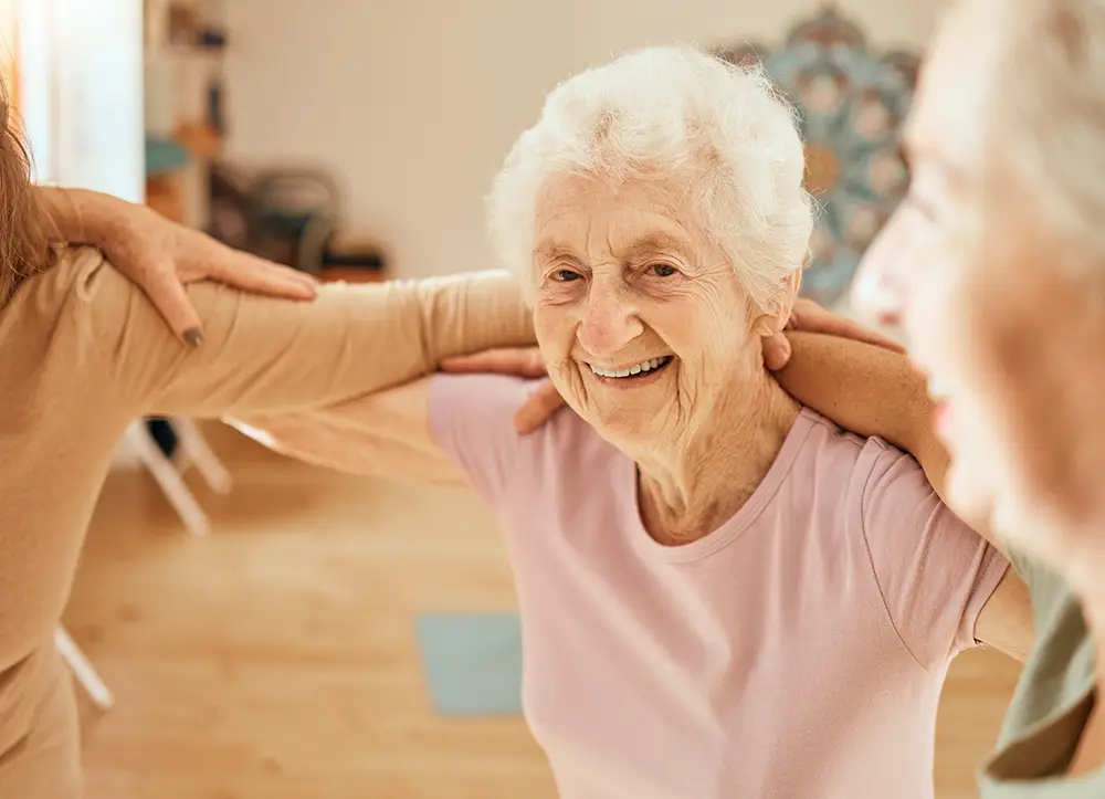 An elderly woman in a light purple shirt smiles as she stands with others, linking arms in a bright indoor setting.